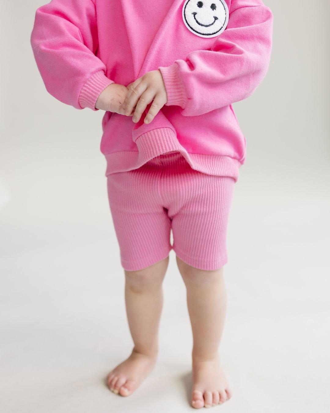 Close up detailed body shot of a toddler girl standing up wearing a bright pink cotton Lucky Panda Kids "Biker Set" which includes ribbed cotton shorts and a sweatshirt with a fuzzy white smiley face emblem on the top right.