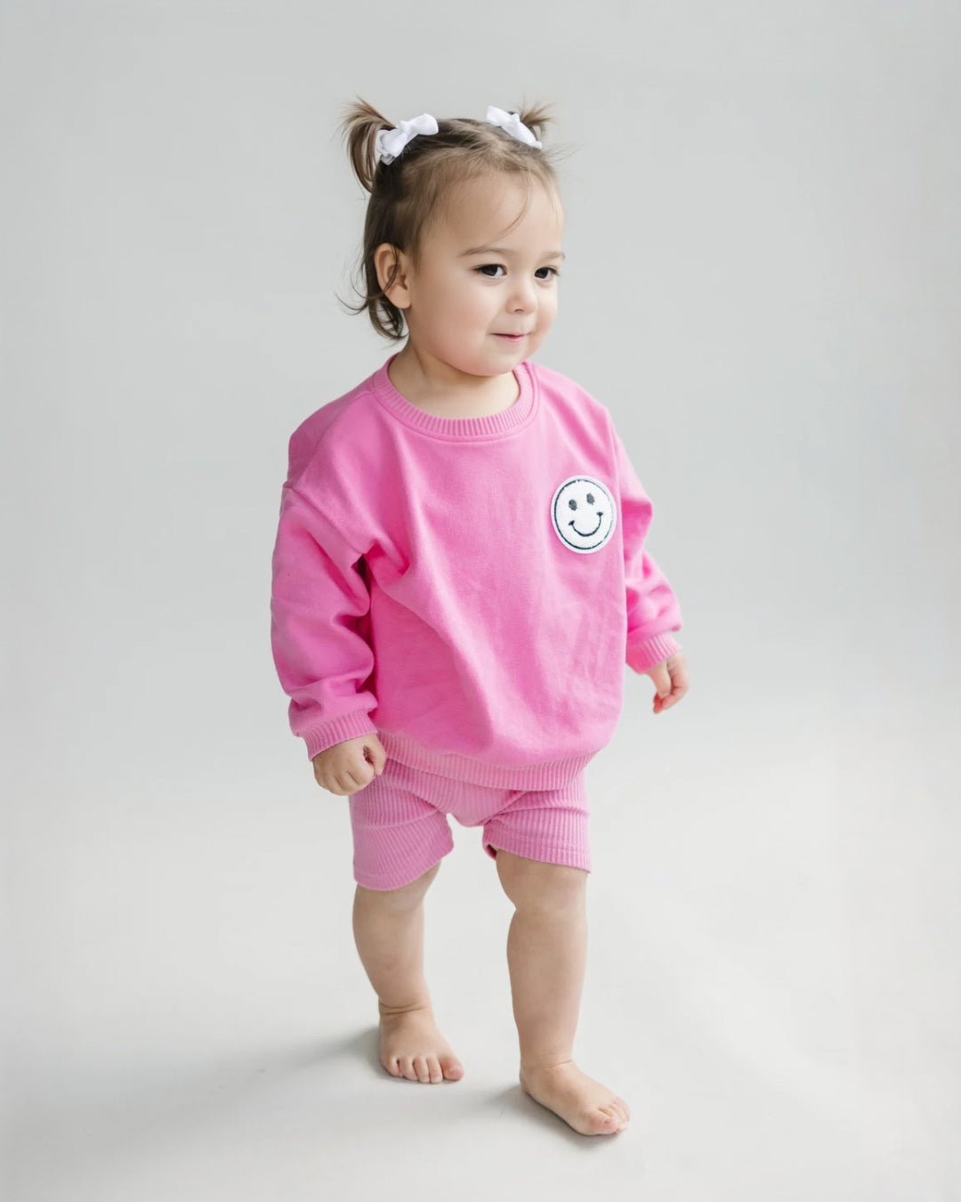 A toddler girl standing up wearing a bright pink cotton Lucky Panda Kids "Biker Set" which includes ribbed cotton shorts and a sweatshirt with a fuzzy white smiley face emblem on the top right.