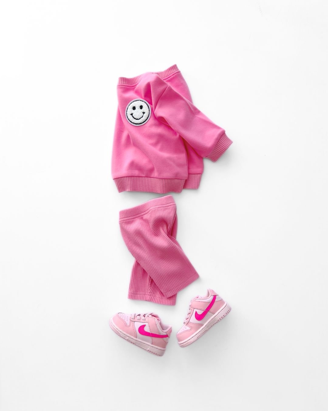 A white background featuring a bright pink cotton Lucky Panda Kids "Biker Set" which includes ribbed cotton shorts and a sweatshirt with a fuzzy white smiley face emblem on the top right.