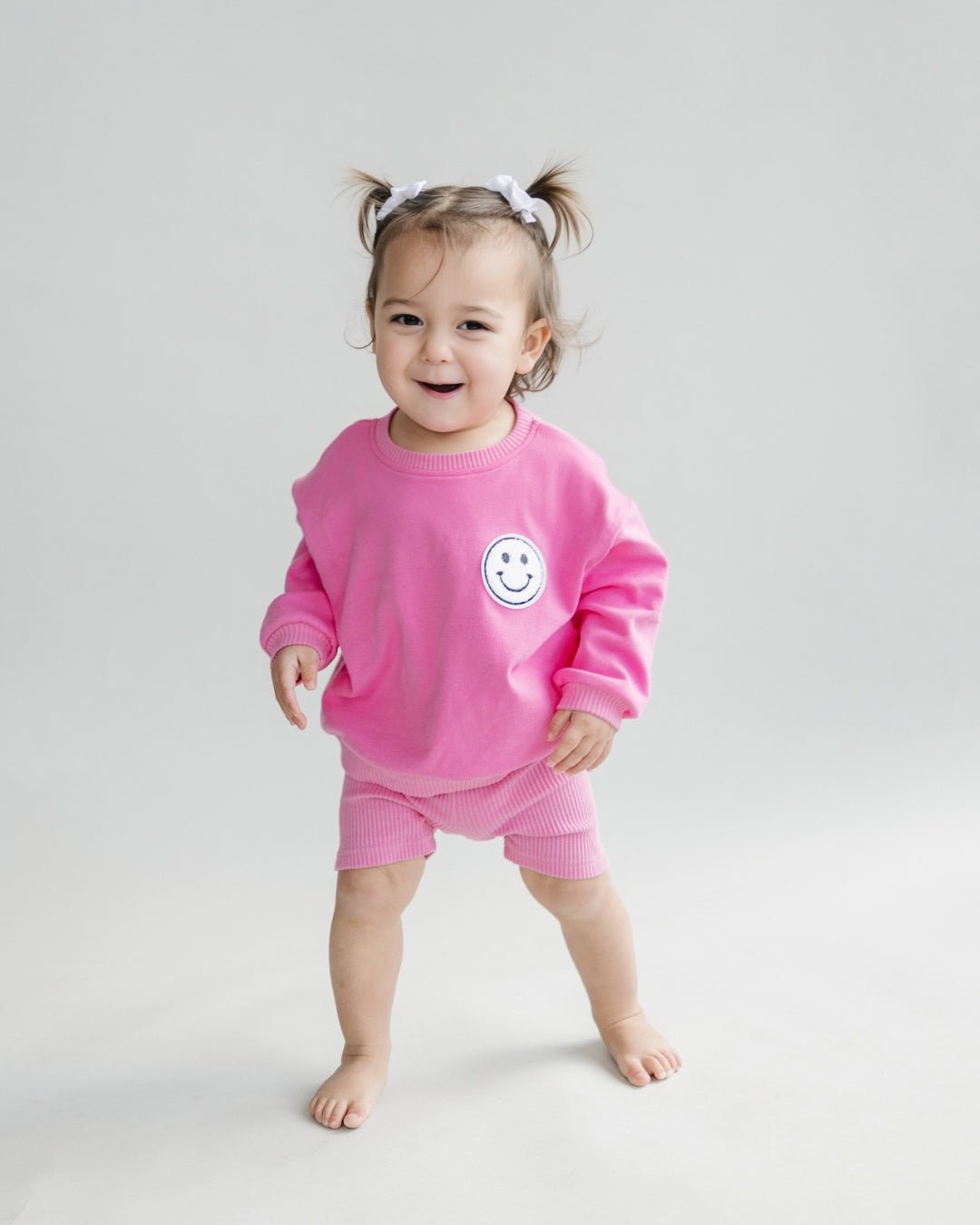 A toddler girl standing up wearing a bright pink cotton Lucky Panda Kids "Biker Set" which includes ribbed cotton shorts and a sweatshirt with a fuzzy white smiley face emblem on the top right.