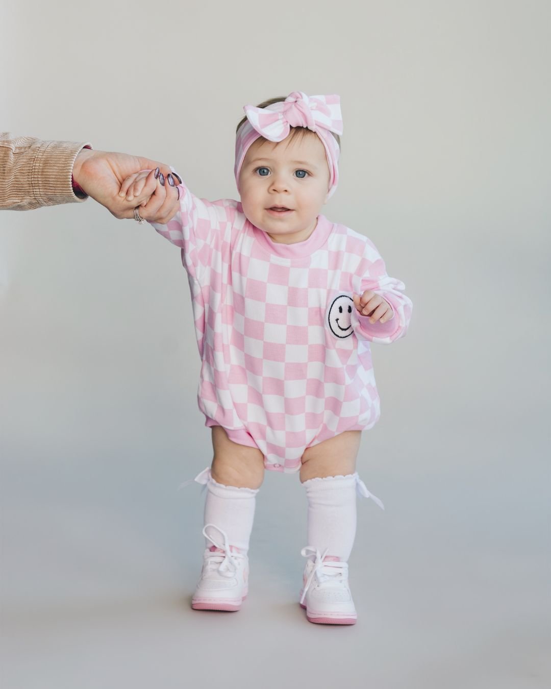 Baby girl standing up wearing a long sleeve light pink and white checkered cotton romper from Lucky Panda Kids. It has a white fuzzy smiley face on the top right corner. She is accessorized with a matching headband and pink shoes and socks.