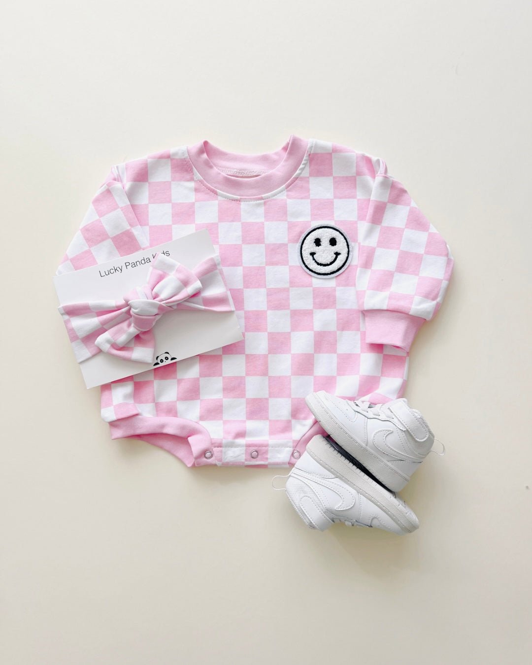 A long sleeve light pink and white checkered cotton romper from Lucky Panda Kids. It has a white fuzzy smiley face on the top right corner. It is accessorized with white nike tennis shoes and a matching headband.