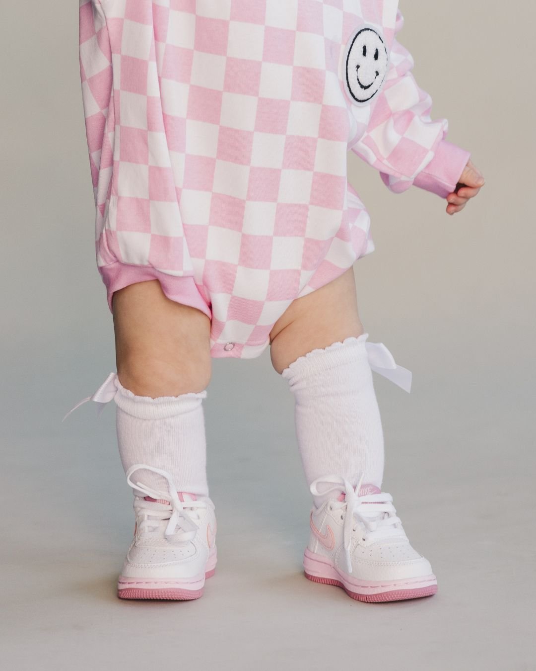 Close up detail below the head of Baby girl standing up wearing a long sleeve light pink and white checkered cotton romper from Lucky Panda Kids. It has a white fuzzy smiley face on the top right corner. She is accessorized with pink shoes and socks.