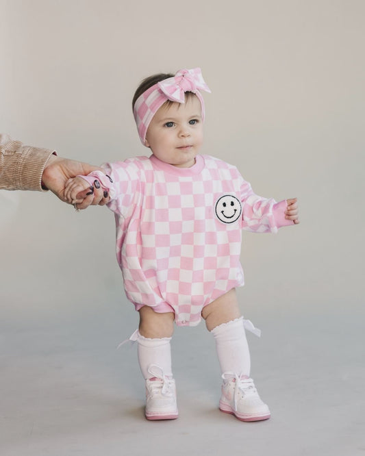 Baby girl standing up wearing a long sleeve light pink and white checkered cotton romper from Lucky Panda Kids. It has a white fuzzy smiley face on the top right corner. She is accessorized with a matching headband and pink shoes and socks.