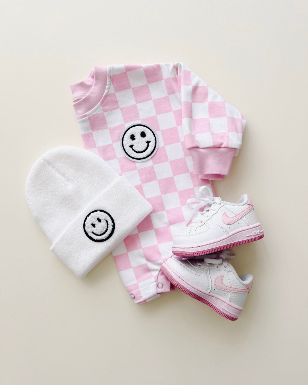 A long sleeve light pink and white checkered cotton romper from Lucky Panda Kids. It has a white fuzzy smiley face on the top right corner. It is accessorized with pink nike tennis shoes and a white smiley face beanie.