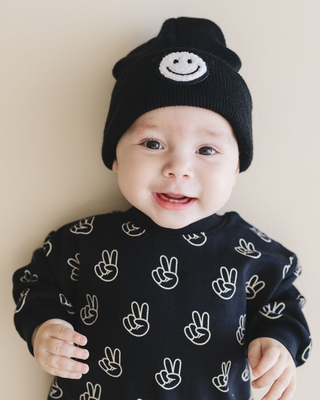 Infant baby boy laying down wearing the long sleeve cotton black bubble romper from Lucky Panda Kids with white peace signs printed on it. 