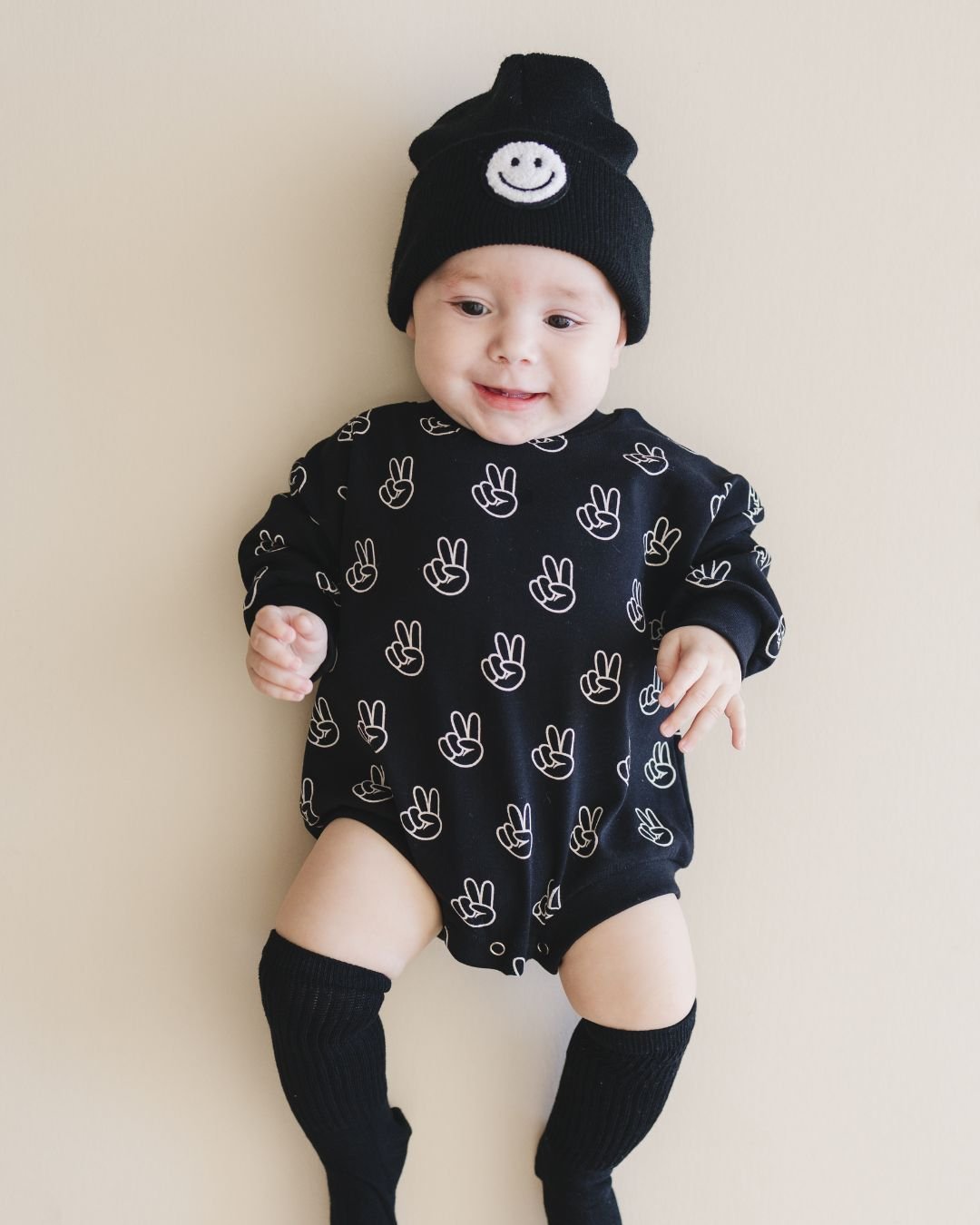 Infant baby boy laying down wearing the long sleeve cotton black bubble romper from Lucky Panda Kids with white peace signs printed on it. 