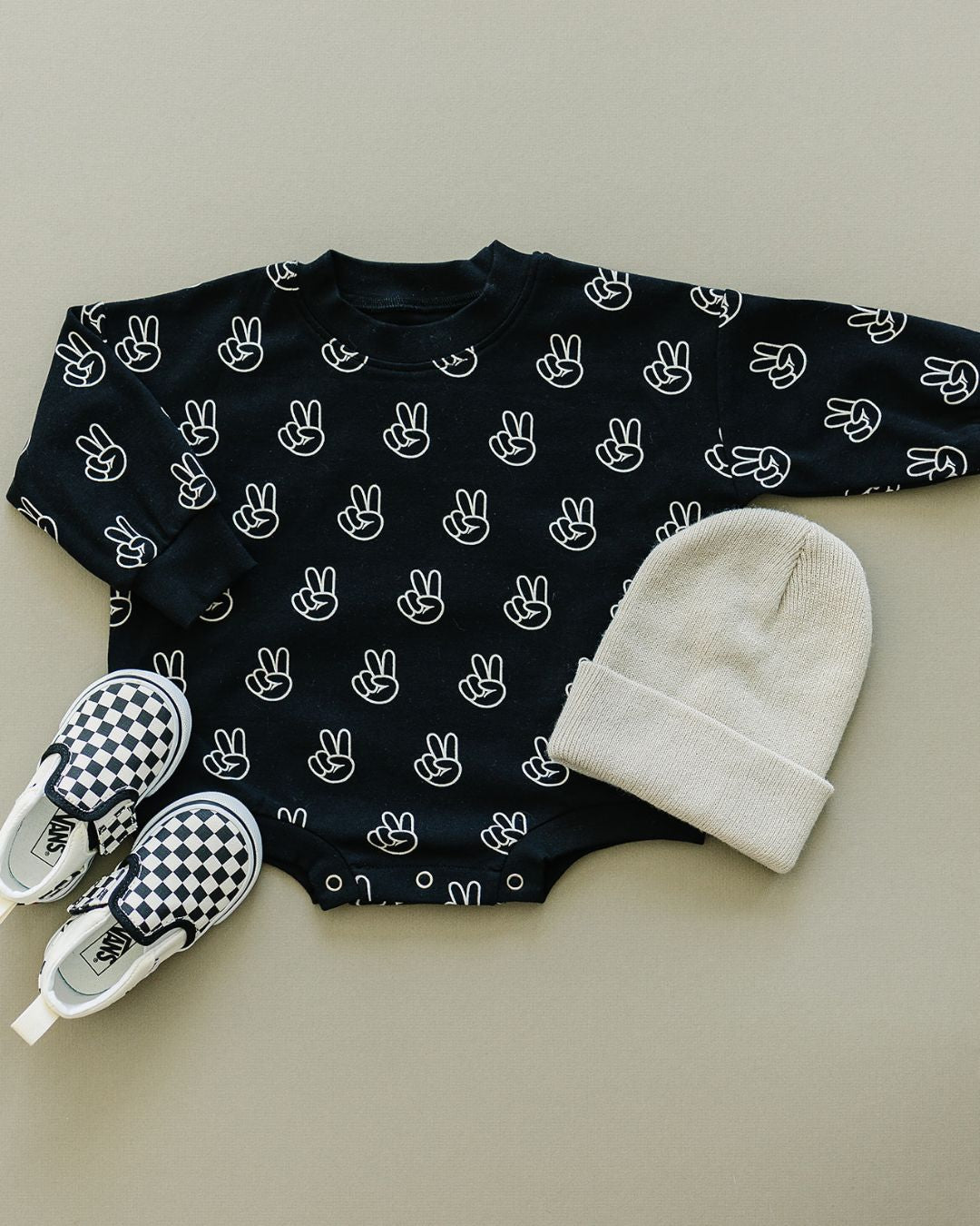 Tan background featuring the long sleeve cotton black bubble romper from Lucky Panda Kids with white peace signs printed on it. It is accessorized with a cream beanie and vans checkered shoes for babies.