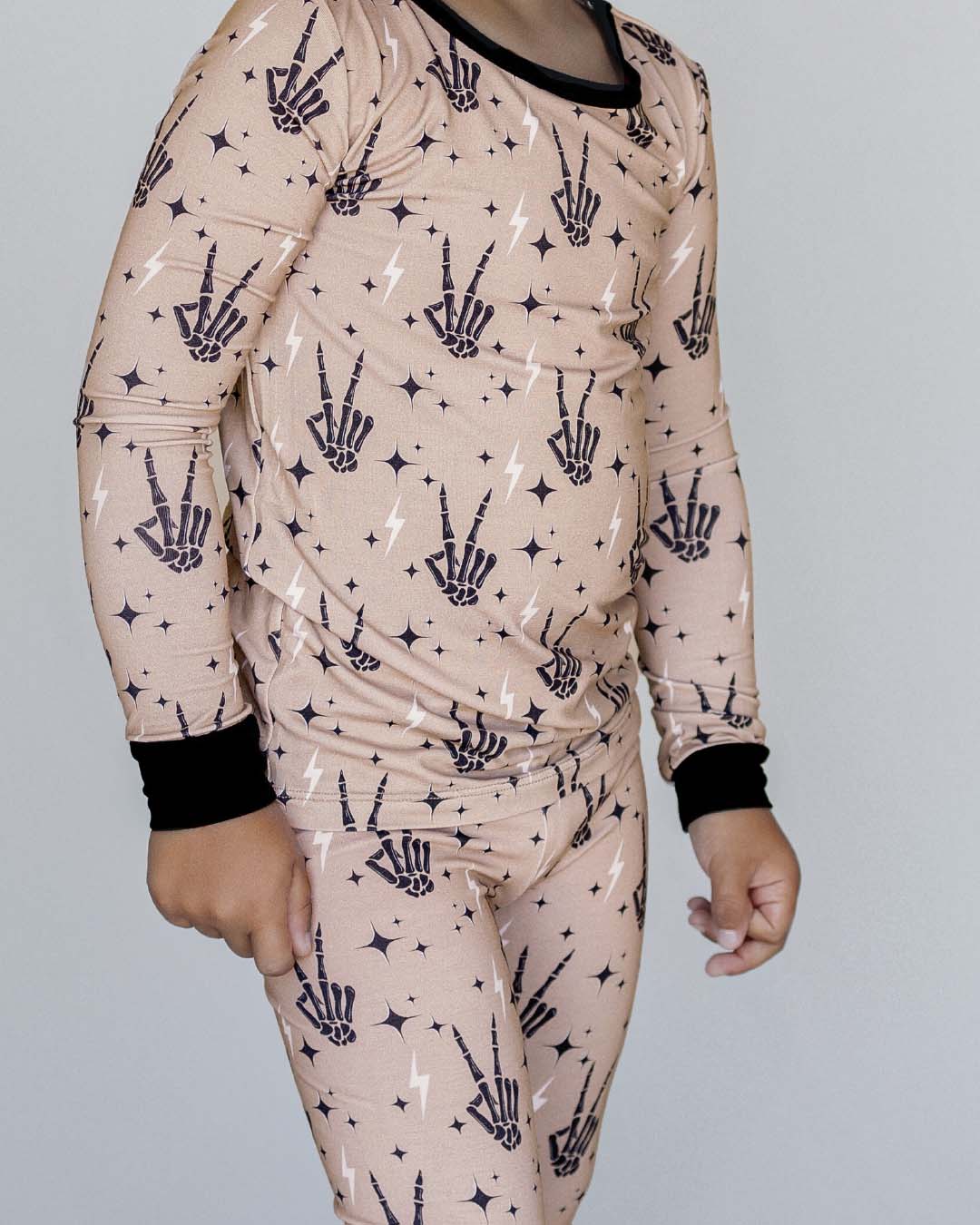 Close up shot of the midsection of a young boy wearing the Lucky Panda Kids viscose bamboo two-piece pajama set in tan designed with black skeleton peace sign hands, white lightning bolts, and black stars.