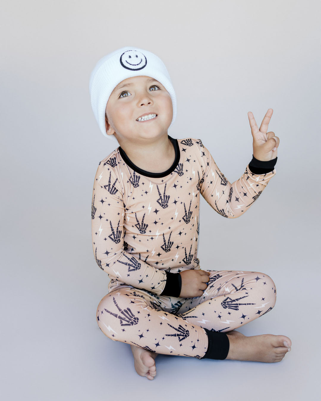 Young boy sitting and holding up a peace sign. He is wearing the Lucky Panda Kids viscose bamboo two-piece pajama set in tan designed with black skeleton peace sign hands, white lightning bolts, and black stars.