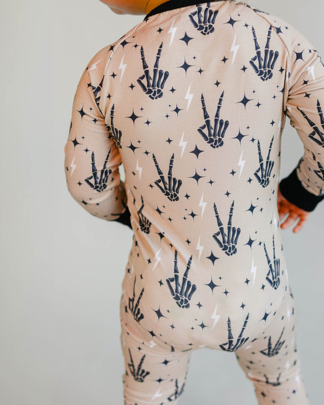 A backside headless shot of a toddler boy standing up wearing tan colored Lucky Panda Kids viscose bamboo zip up pajamas in a halloween style with printed black skeleton peace signs, white lightning bolts, and black stars. 