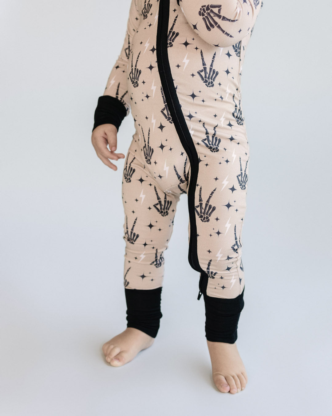 Below the head shot of a toddler boy standing up wearing tan colored Lucky Panda Kids viscose bamboo zip up pajamas in a halloween style with printed black skeleton peace signs, white lightning bolts, and black stars. 