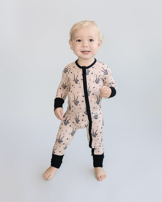 A toddler boy standing up wearing tan colored Lucky Panda Kids viscose bamboo zip up pajamas in a halloween style with printed black skeleton peace signs, white lightning bolts, and black stars. 