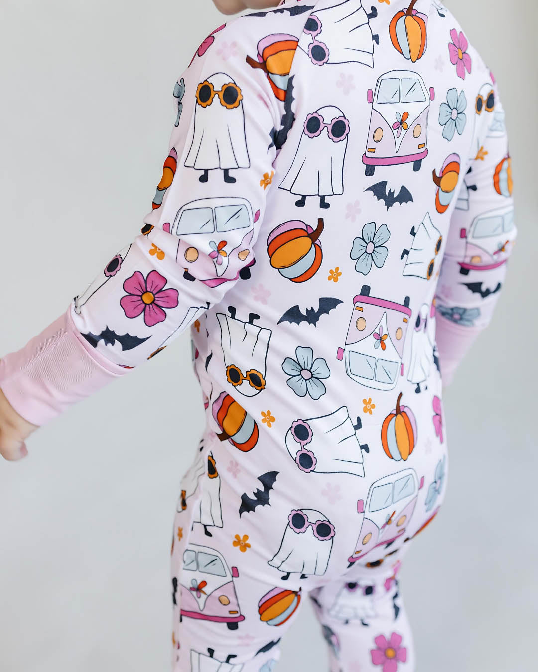 Close up body shot of a toddler girl standing with her back to the camera. She is wearing a light pink Lucky Panda Kids zip up pajama in the style of halloween. It has white ghosts, multicolored orange pumpkins, blue and pink flowers, and black bats printed on it.