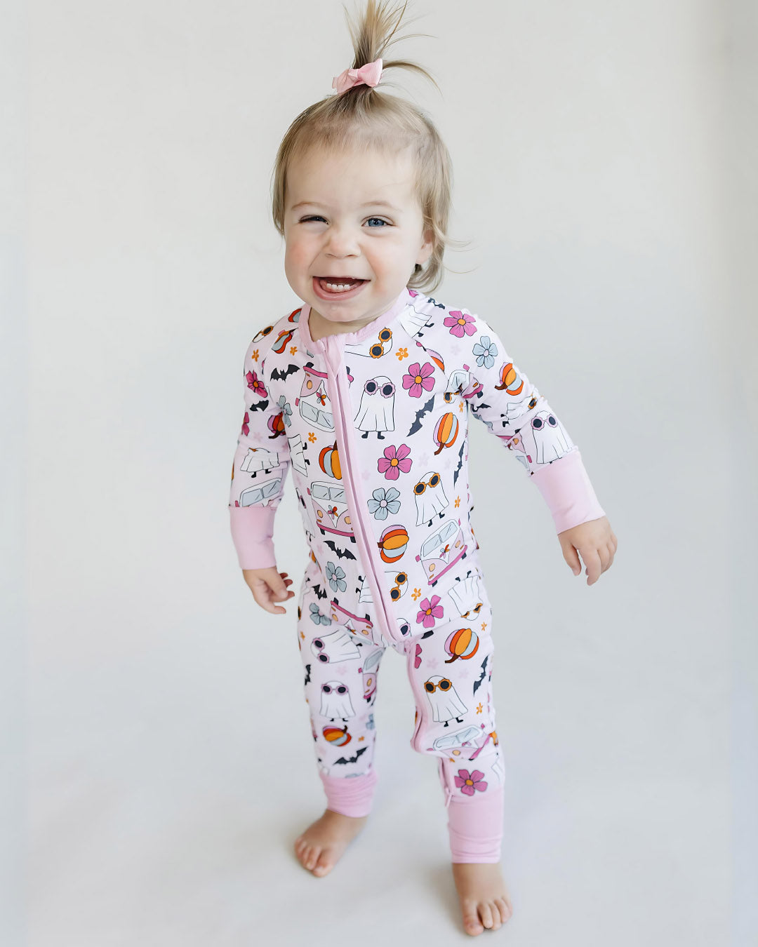 A toddler girl standing up smiling. She is wearing a light pink Lucky Panda Kids zip up pajama in the style of halloween. It has white ghosts, multicolored orange pumpkins, blue and pink flowers, and black bats printed on it.