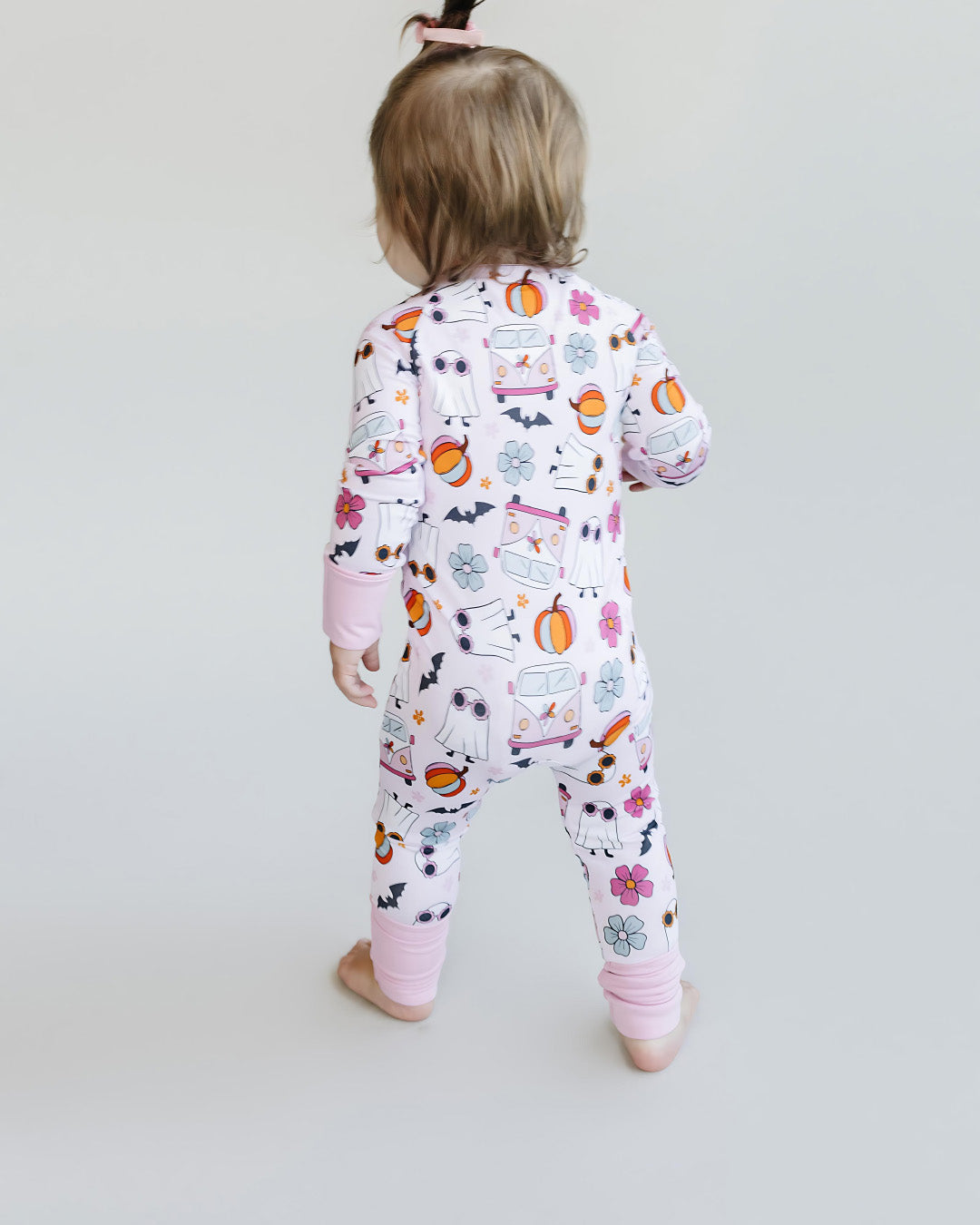 A toddler girl standing with her back to the camera. She is wearing a light pink Lucky Panda Kids zip up pajama in the style of halloween. It has white ghosts, pink hippie vans, multicolored orange pumpkins, blue and pink flowers, and black bats printed on it.