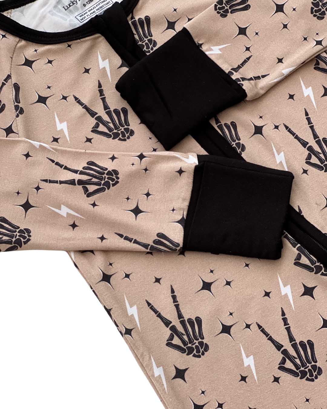 Detailed close up of a tan colored Lucky Panda Kids viscose bamboo zip up pajamas in a halloween style with printed black skeleton peace signs, white lightning bolts, and black stars. 