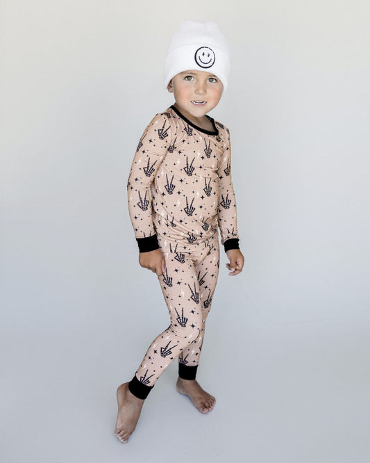 Young boy standing up wearing Lucky Panda Kids viscose bamboo two-piece pajama set in tan designed with black skeleton peace sign hands, white lightning bolts, and black stars.